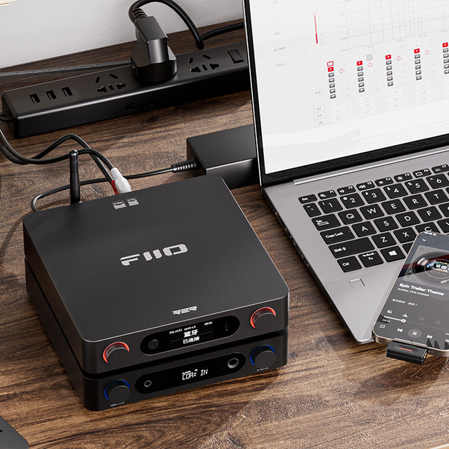 FiiO B15 R2R שני מכשירי אודיו שולחניים מונחים זה על גבי זה ומונחים על שולחן עבודה לצד מחשב לפטום, טלפון נייד, מטעני חשמל. המכשיר העליון הוא הבי.אר. 15 והתחתון הוא מגבר אוזניות של פיו