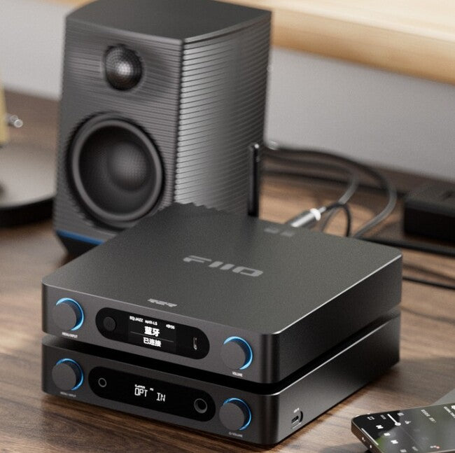 FiiO BR15 R2R שני מכשירי אודיו שולחניים שחורים המונחים זה על גבי זה על שולחן עבודה בצבע אגוז ומאחוריהם על גבי השולחן רמקול שחור מוגבר של פיו