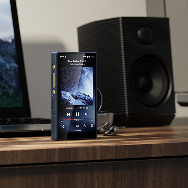 נגן אודיו דיגטלי fiio m21 dapמונח אנכית על שולחן עבודה מאחוריו מחשב לפטופ לצידו אוזניו אינאיר אלחוטיות של פיו ורמקול שולחני של פיו