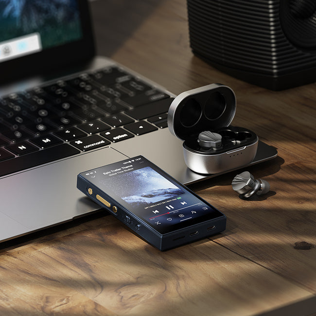 נגן אודיו דיגטלי FIIO M21 DAPבצבע שחור מונח בשכיבה על שולחן עבודה כשהחלק העליון של הנגן מונח על מחשב אישי לפטופ פתוח ולצידו אוזניות אלחוטיות של פיו, ברקע מאחרו רמקול שולחני שחור של פיו